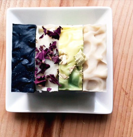 Four artisanal handmade soaps arranged side-by-side on a white square plate, set on a wooden table. The soaps are colored black, white with dried purple flowers, pale yellow with dried green flowers, and cream/beige with dried flowers.