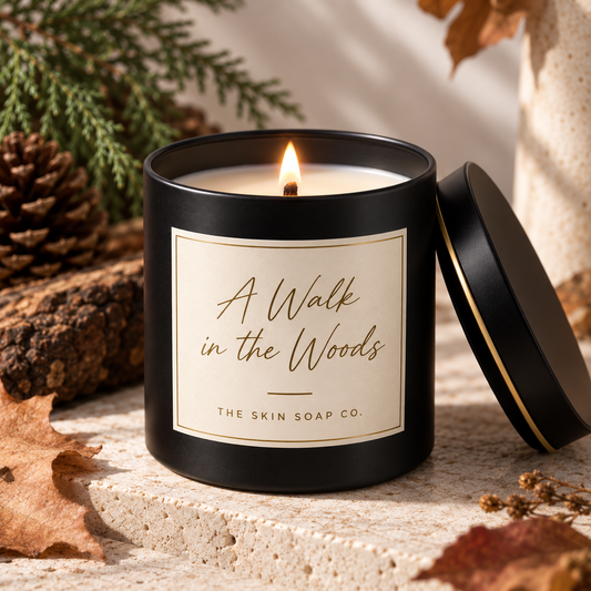A black candle tin with a lit wick and cream-colored wax, labeled 'A Walk in the Woods' by 'The Skin Soap Co.', styled on a stone surface with autumn leaves, pinecones, and evergreen branches.