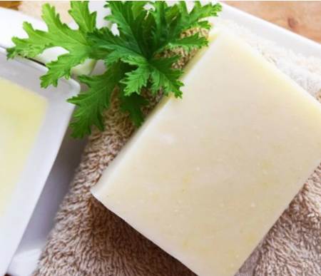 A rectangular bar of pale creamy soap resting on a brown towel, garnished with a sprig of fresh green parsley, with a second soap bar partially visible in a white dish in the background.