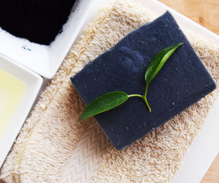 A rectangular dark charcoal soap bar with two green leaves on top, resting on a beige textured towel over a white tray.