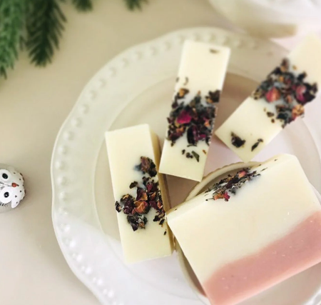 Four rectangular bars of handmade soap on a white plate, featuring cream-colored top layers, a soft pink bottom layer on one bar, and dried flower petals on top, set against a warm neutral background with a small decorative owl figurine.